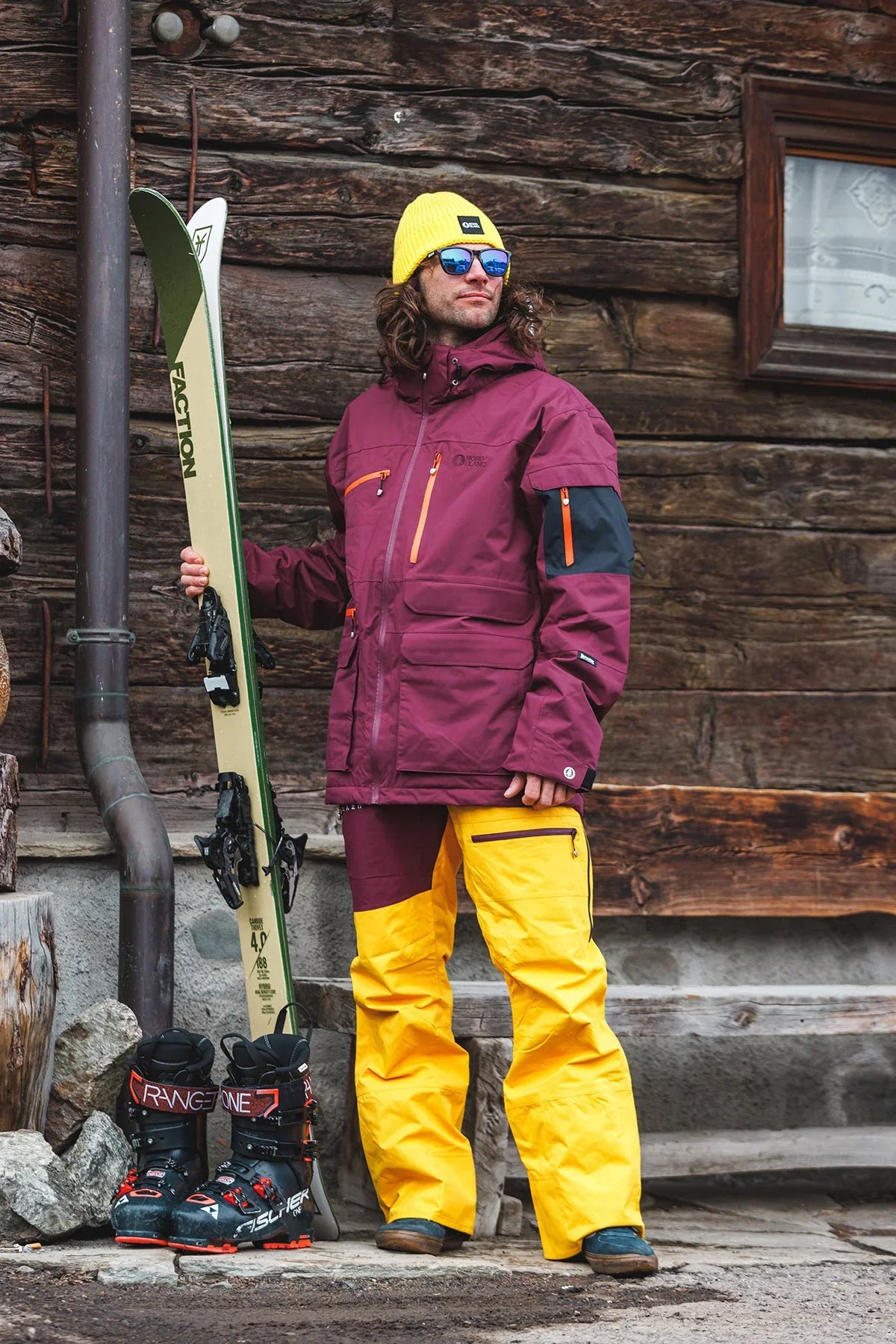 Skier in yellow ski trousers and a burgundy jacket by a wooden cabin
